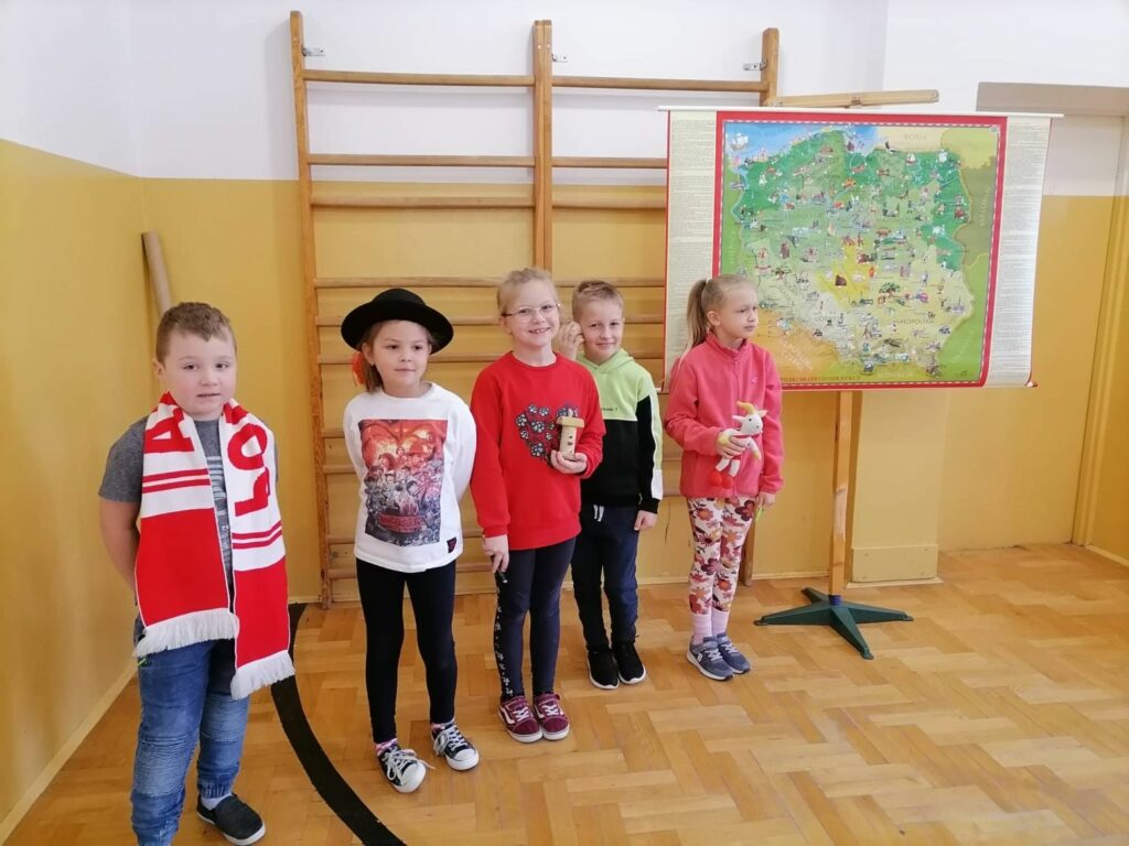 Pod ścianą na specjalnym wieszaku wisi wielka kolorowa mapa Polski, obok której stoi dwóch chłopców i trzy dziewczynki. Każde z dzieci ma na sobie lub w rękach jakiś gadżet np. góralski kapelusz, muszlę z Bałtyku, mały ulik z Kamiannej, czy biało czerwony szalik.