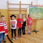 Pod ścianą na specjalnym wieszaku wisi wielka kolorowa mapa Polski, obok której stoi dwóch chłopców i trzy dziewczynki. Każde z dzieci ma na sobie lub w rękach jakiś gadżet np. góralski kapelusz, muszlę z Bałtyku, mały ulik z Kamiannej, czy biało czerwony szalik.
