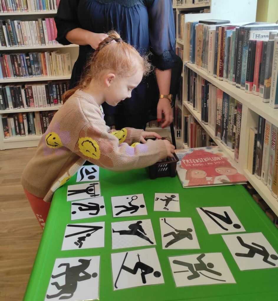 na zdjęciu widać dziewczynkę stojąca przed zielonym stolikiem na którym poukładano czarno-białe obrazki.