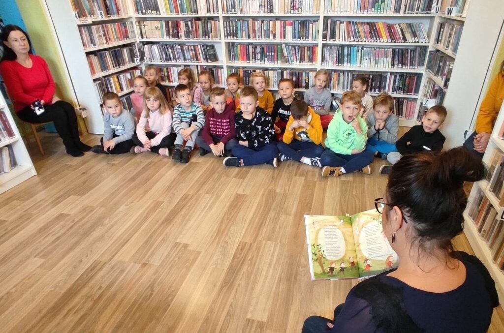 na zdjęciu widać dzieci siedzące na podłodze oraz bibliotekarkę czytającą im książkę.