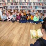 na zdjęciu widać dzieci siedzące na podłodze oraz bibliotekarkę czytającą im książkę.