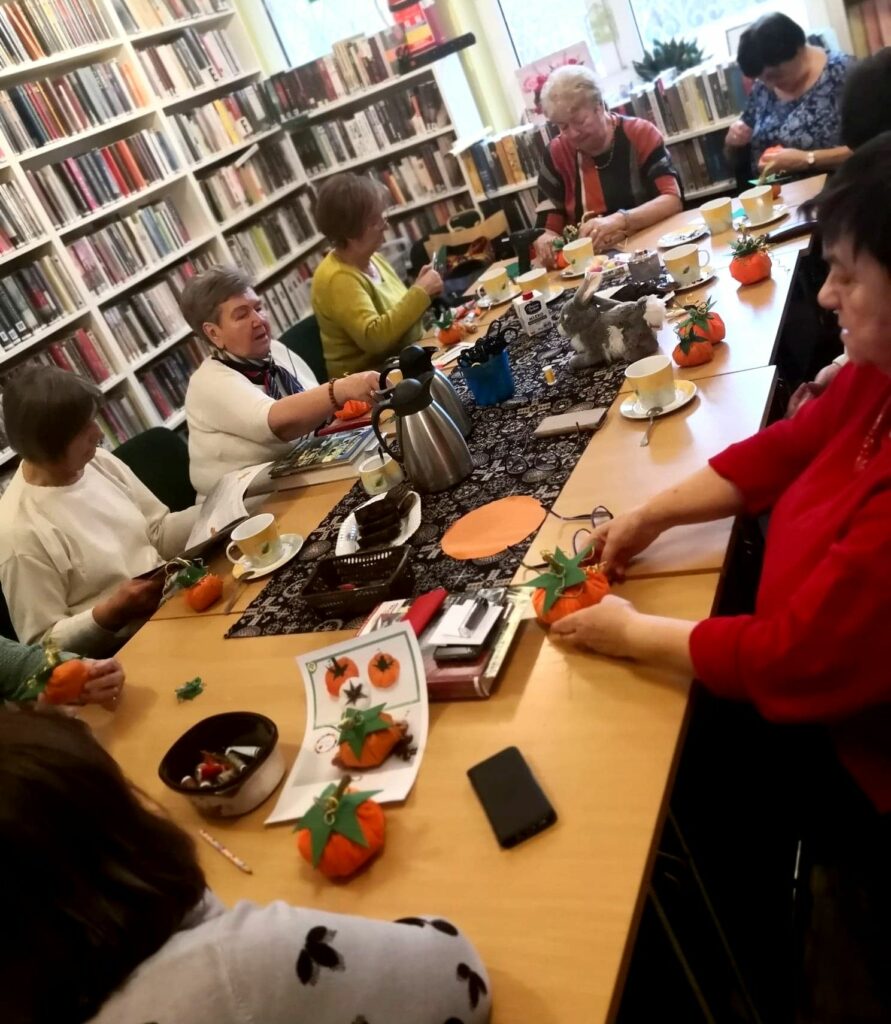 Na zdjęciu widać seniorki siedzące wokół stołu w sali bibliotecznej