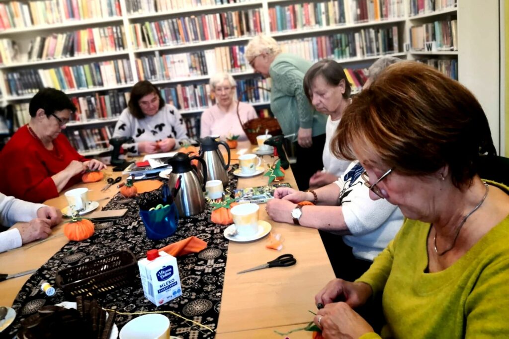 Na zdjęciu widać seniorki siedzące wokół stołu w sali bibliotecznej