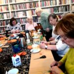 Na zdjęciu widać seniorki siedzące wokół stołu w sali bibliotecznej