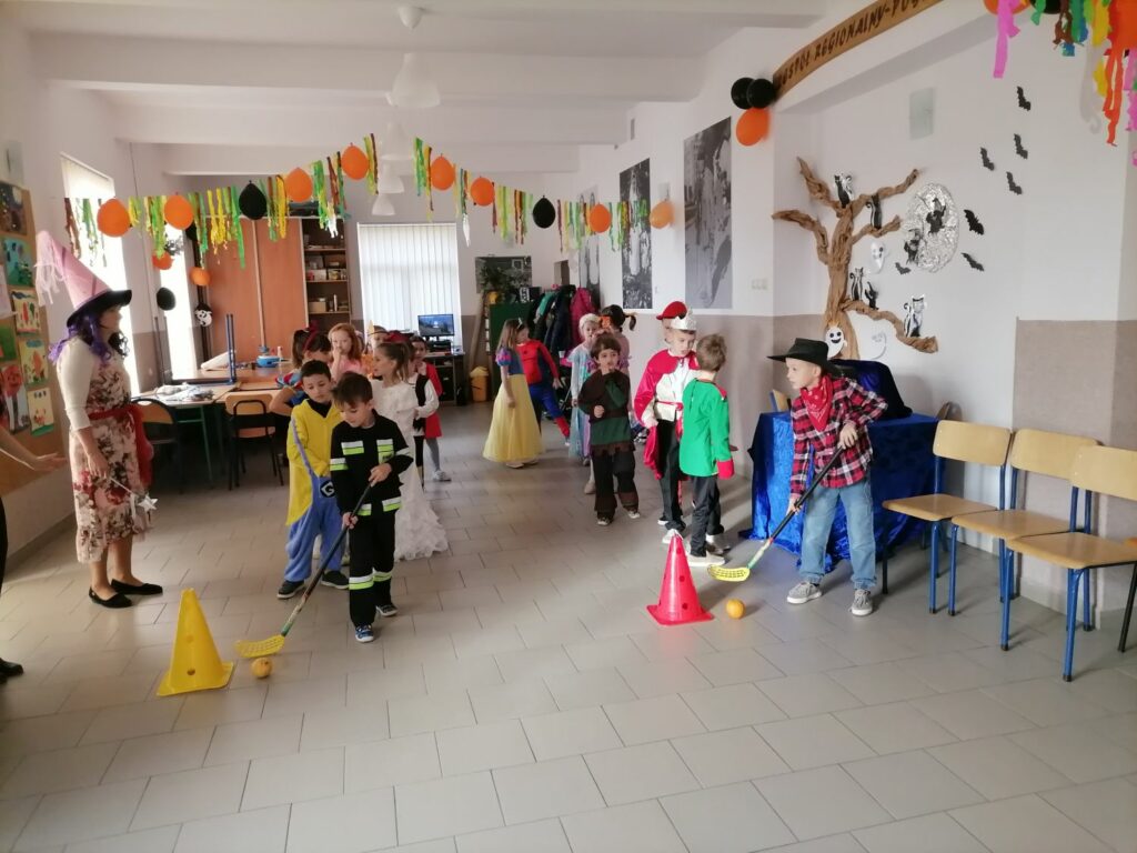 W dużej sali dzieci stoją w dwuch rzędach i rywalizują ze sobą w konkursie. Z boku stoi pani bibliotekarka i kibicuje dzieciom. Dzieci poprzebierane sa w kolorowe stroje.