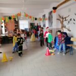 W dużej sali dzieci stoją w dwuch rzędach i rywalizują ze sobą w konkursie. Z boku stoi pani bibliotekarka i kibicuje dzieciom. Dzieci poprzebierane sa w kolorowe stroje.