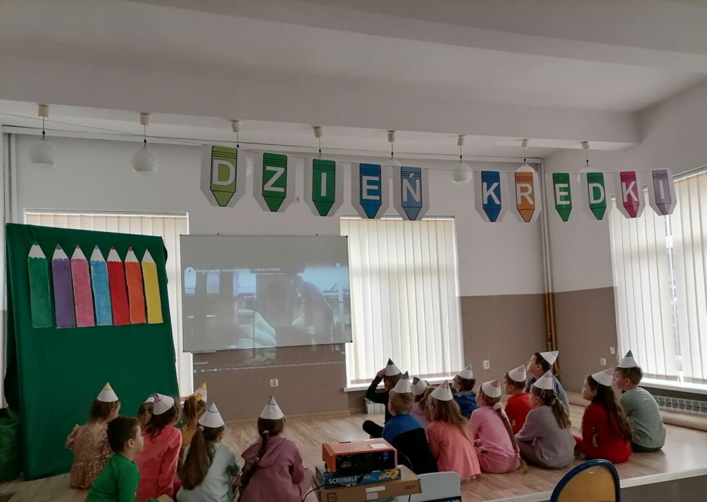 Dzieci w kolorowych czapeczkach siedzą na ziemi przodem do ekranu i oglądają bajkę