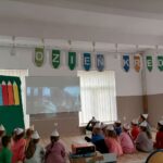 Dzieci w kolorowych czapeczkach siedzą na ziemi przodem do ekranu i oglądają bajkę