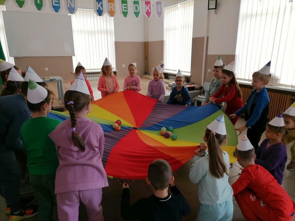 Dzieci kolorowo ubrane w czapeczkach stoją wokół chusty klanza na której turlają się małe kolorowe kuleczki