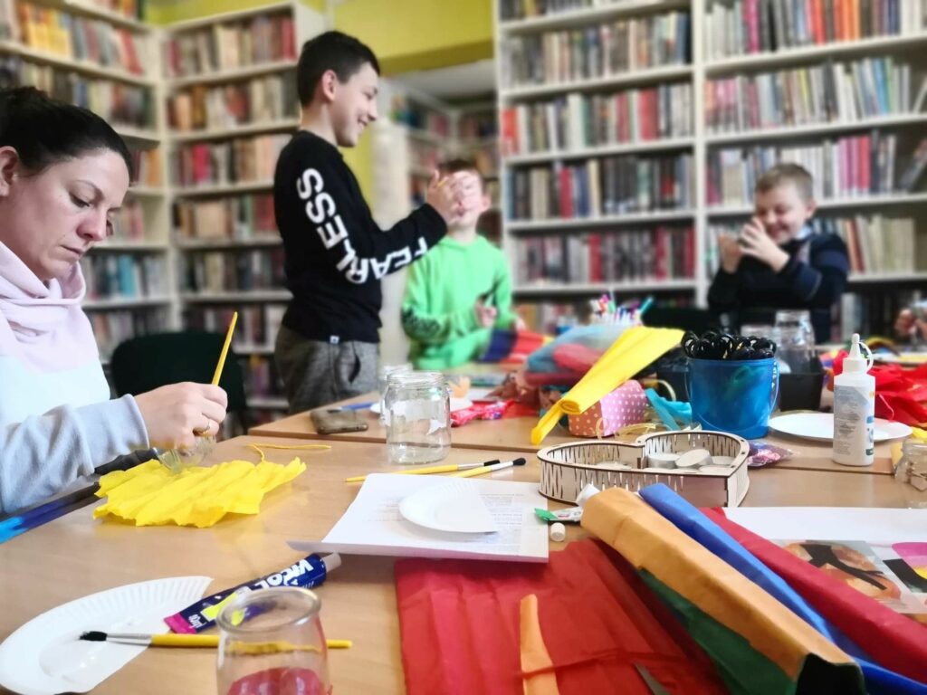 na zdjęciu widać dzieci siedzące przy stole na którym jest wiele materiałów plastycznych takich jak np. kleje, bibuła, kolorowy papier.
