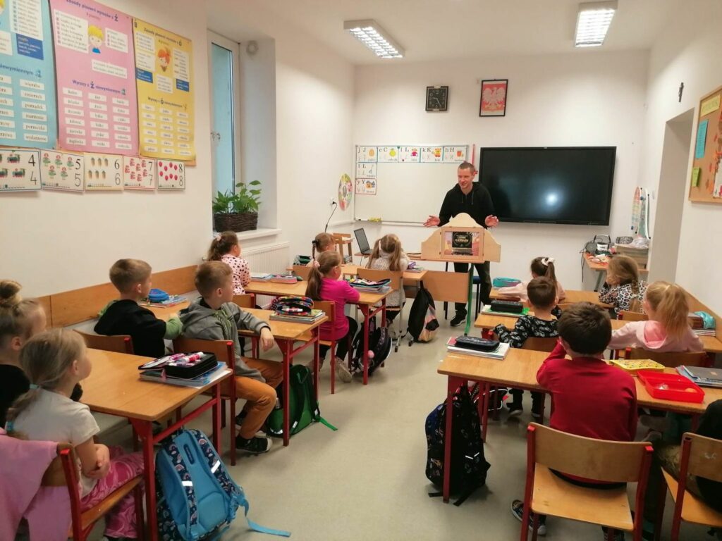 W szkolnej klasie siedzący w ławkach uczniowie oglądają prezentowany im przez bibliotekarza teatrzyk obrazkowy Kamishibai.
