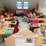 Grupa uczniów siedzi w ławkach, a także na podłodze pod oknami i z uwagą oglądają prezentowaną w teatrzyku kamishibai „Legendę o Skarbniku”.