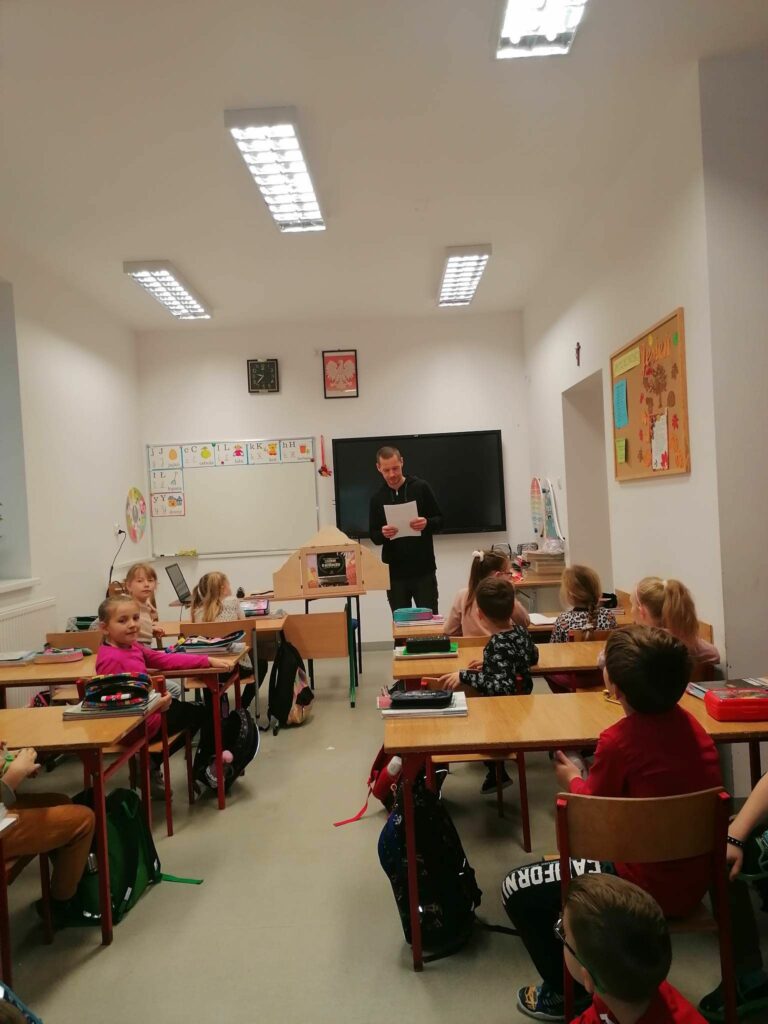 Pan bibliotekarz stoi na środku klasy i czyta słuchającym go z rozbawieniem uczniom zabawne śląskie zwroty i słowa gwarowe.