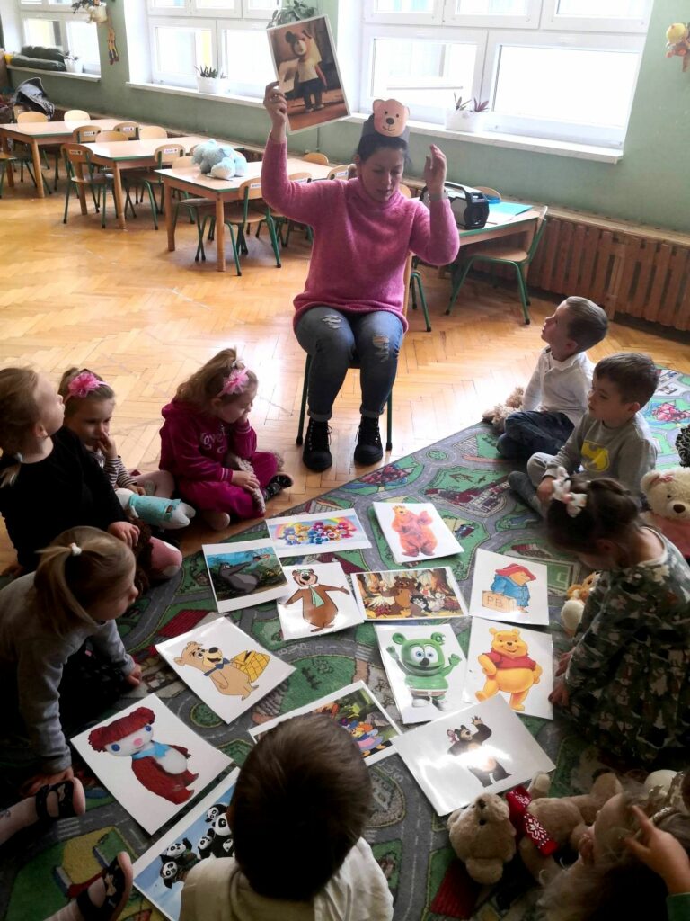 na zdjęciu widać dzieci siedzące na dywanie oraz bibliotekarkę okazujące im ilustracje przedstawiające misie z bajek