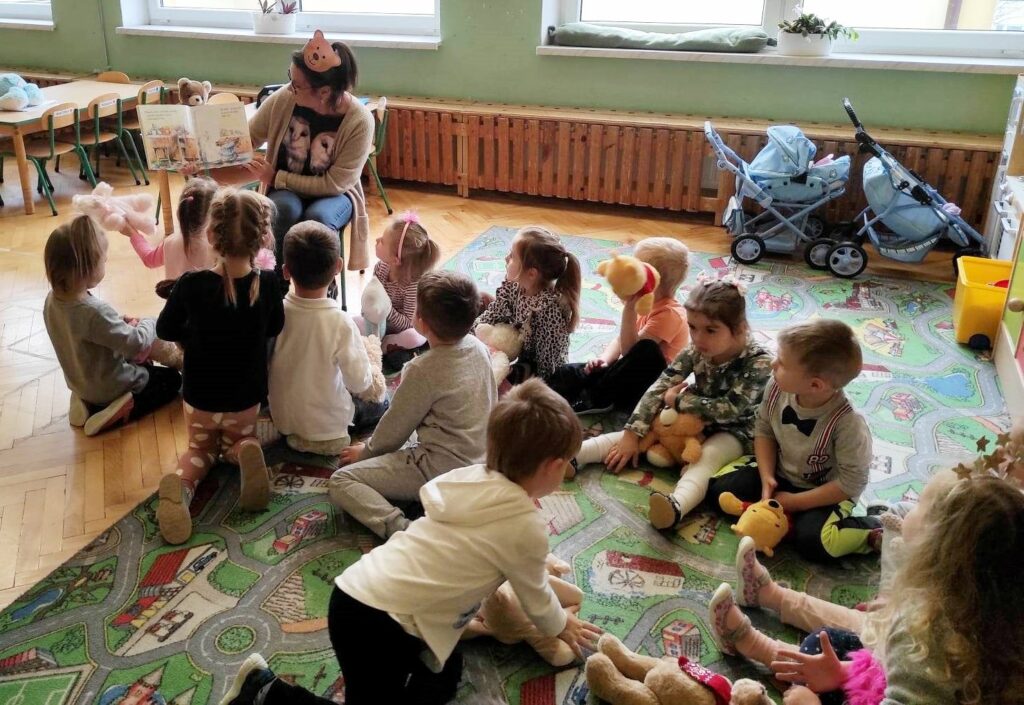 na zdjęciu widać dzieci siedzące na dywanie i słuchające czatającą im bibliotekarkę