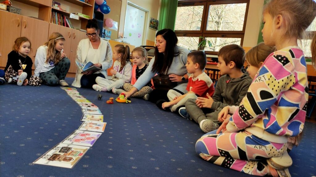 na zdjęciu widać dzieci i dwie bibliotekarki siedzące na granatowej wykładzinie