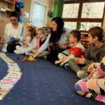 na zdjęciu widać dzieci i dwie bibliotekarki siedzące na granatowej wykładzinie