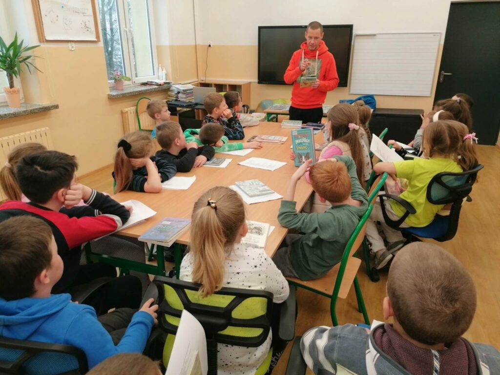 W szkolnej sali grupa uczniów siedzi wokół złączonych ze sobą stolików i słucha stojącego przed nimi pana bibliotekarza, który czyta im książkę o piłce nożnej.