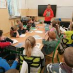 W szkolnej sali grupa uczniów siedzi wokół złączonych ze sobą stolików i słucha stojącego przed nimi pana bibliotekarza, który czyta im książkę o piłce nożnej.