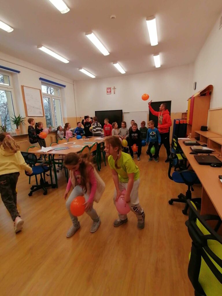 Dzieci zostały podzielone w pary i rozgrywają między sobą wyścigi z balonami trzymanymi w kolanach. Na pierwszym planie widać dwie rywalizujące ze sobą dziewczynki.