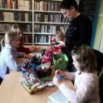 Na zdjęciu widać sale biblioteczną, stół na nim materiały plastyczne a wokół siedzące dzieci oraz stojąca bibliotekarkę.