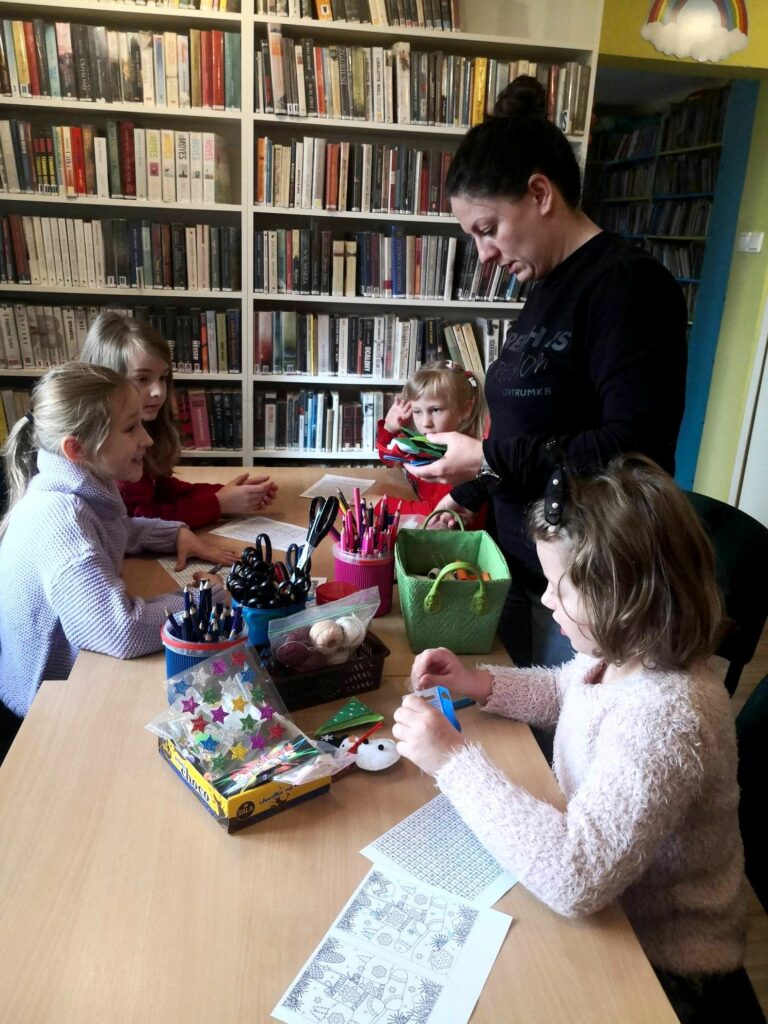 Na zdjęciu widać sale biblioteczną, stół na nim materiały plastyczne a wokół siedzące dzieci oraz stojąca bibliotekarkę.