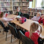 Na zdjęciu widać sale biblioteczną i stół i wokół niego siedzące dzieci i czytającą im bibliotekarkę.