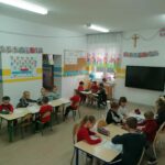 Biorąca udział w zajęciach grupa dzieci siedzi przy stolikach i kredkami koloruje rysunki przedstawiające św. Mikołaja.
