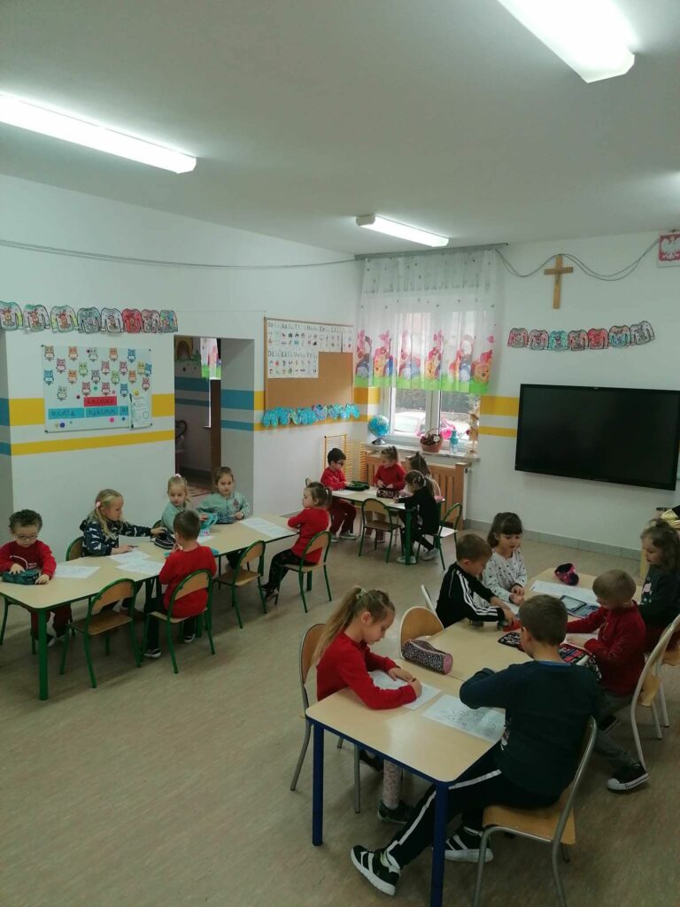 Biorąca udział w zajęciach grupa dzieci siedzi przy stolikach i kredkami koloruje rysunki przedstawiające św. Mikołaja.