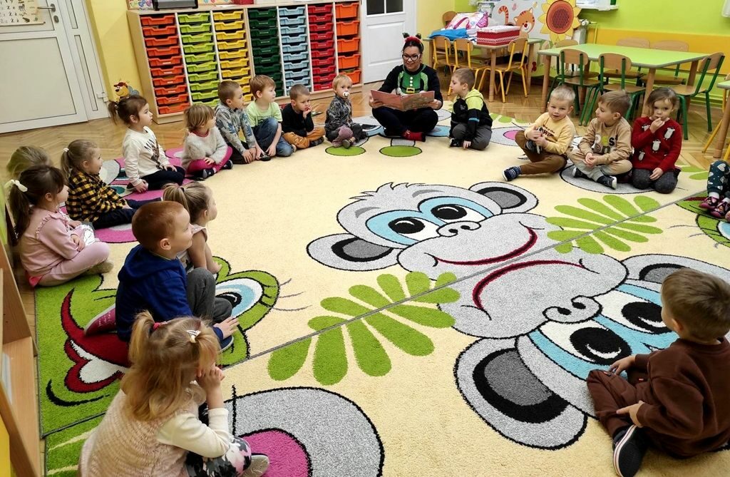 na zdjęciu widać dzieci siedzące na kolorowym dywanie i słuchające książkę czytaną im przez bibliotekarkę
