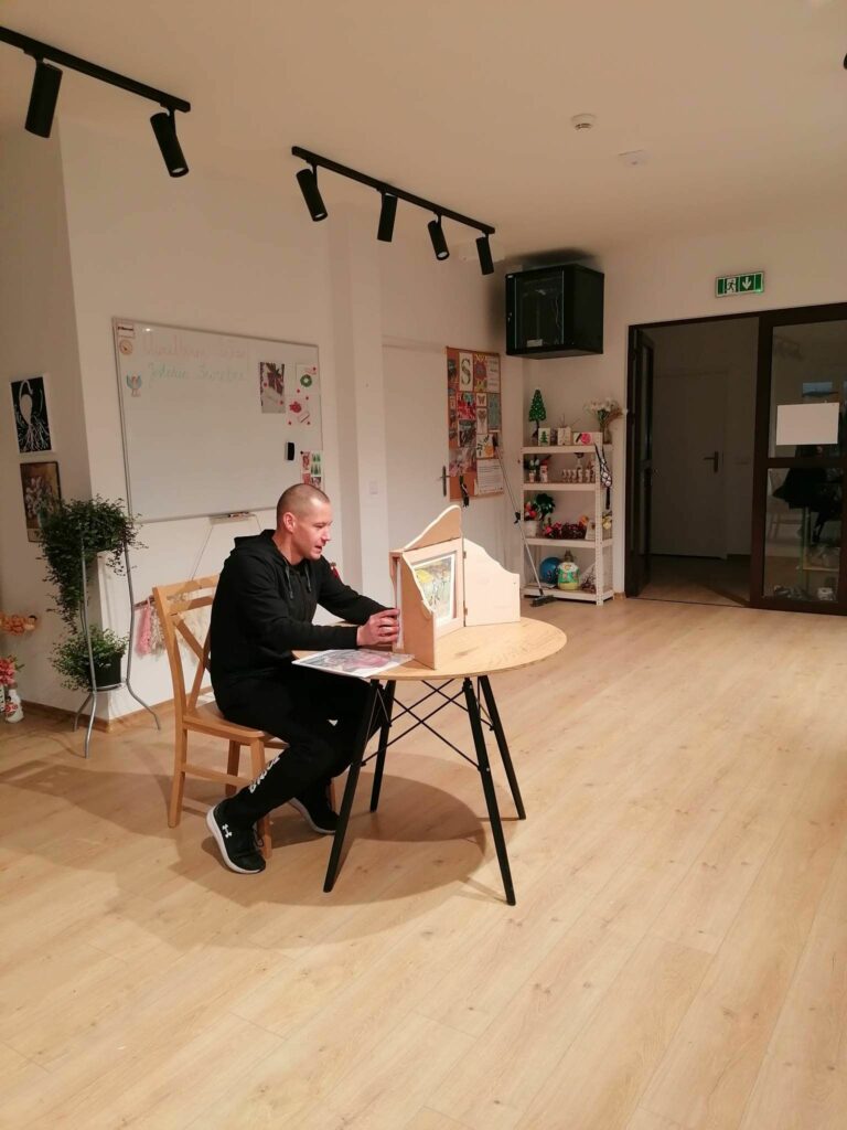 Pan bibliotekarz siedzi przy niewielkim okrągłym stoliku na którym ustawiony jest drewniany teatrzyk kamishibai i czyta przedstawienie pt. „Mój znajomy…” opowiadające o losach Nikifora.