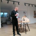Pan bibliotekarz stoi obok stolika na którym znajduje się drewniany teatrzyk kamishibai i w rękach trzyma wydrukowane kolorowe obrazy Nikifora, które za chwilę będą malować Seniorzy.