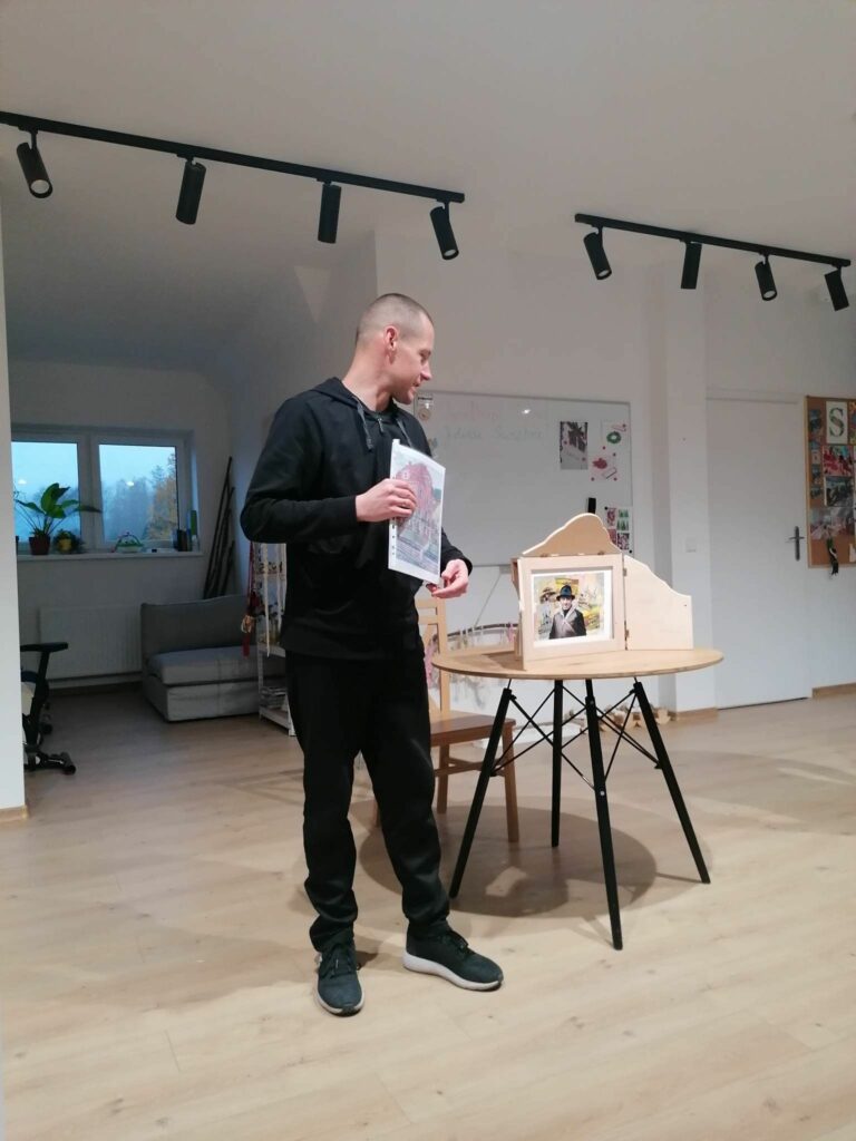 Pan bibliotekarz stoi obok stolika na którym znajduje się drewniany teatrzyk kamishibai i w rękach trzyma wydrukowane kolorowe obrazy Nikifora, które za chwilę będą malować Seniorzy.