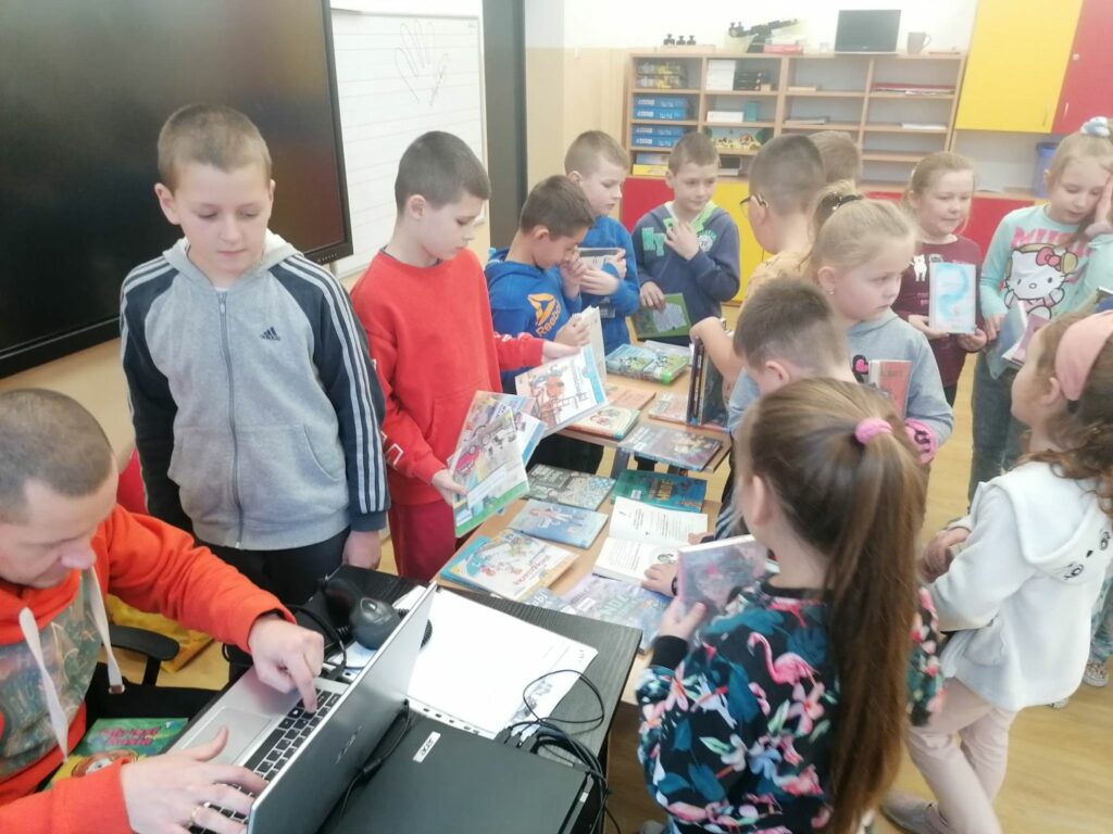 Spora grupa dzieci stoi wokół stolików na których są rozłożone kolorowe książki i wybierają te, które chcą wypożyczyć. W rogu zdjęcia widać siedzącego przy biurku pana bibliotekarza, który przy pomocy laptopa wypożycza lektury