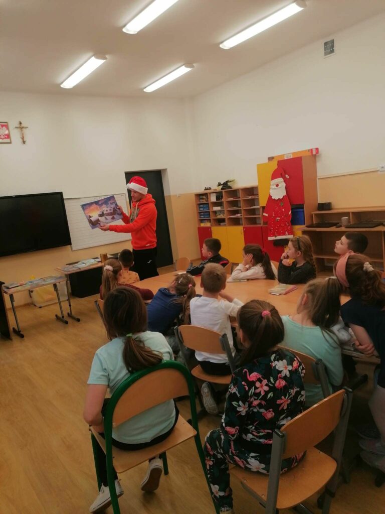 W szkolnej sali grupa uczniów siedzi wokół złączonych ze sobą stolików i słucha stojącego przed nimi pana bibliotekarza, który ubrany w czapkę mikołajową czyta im bajkę pt. „Przyjaciele z piernika”.