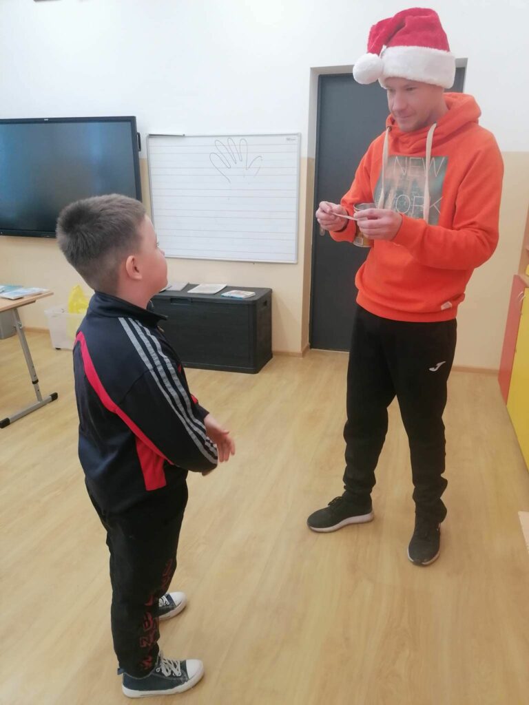 Na zdjęciu widać stojącego pana bibliotekarza z czapką mikołaja na głowie i chłopca, który musi odpowiedzieć na zadaną mu zimową zagadkę. Nagrodą jest słodki cukierek.