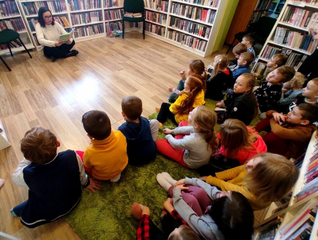 na zdjęciu widać dzieci siedzące na podłodze oraz czytającą im bibliotekarkę