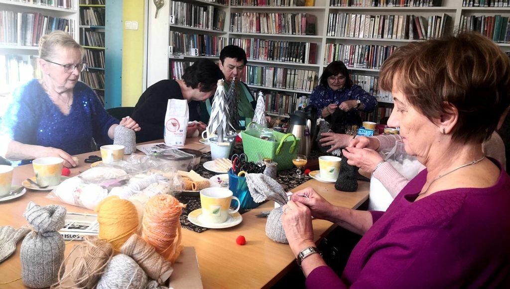 na zdjęciu widać panie siedzące przy stole w sali bibliotecznej. Na stole widać różne materiały do pracy takie jak np. włóczki.