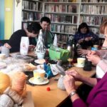 na zdjęciu widać panie siedzące przy stole w sali bibliotecznej. Na stole widać różne materiały do pracy takie jak np. włóczki.