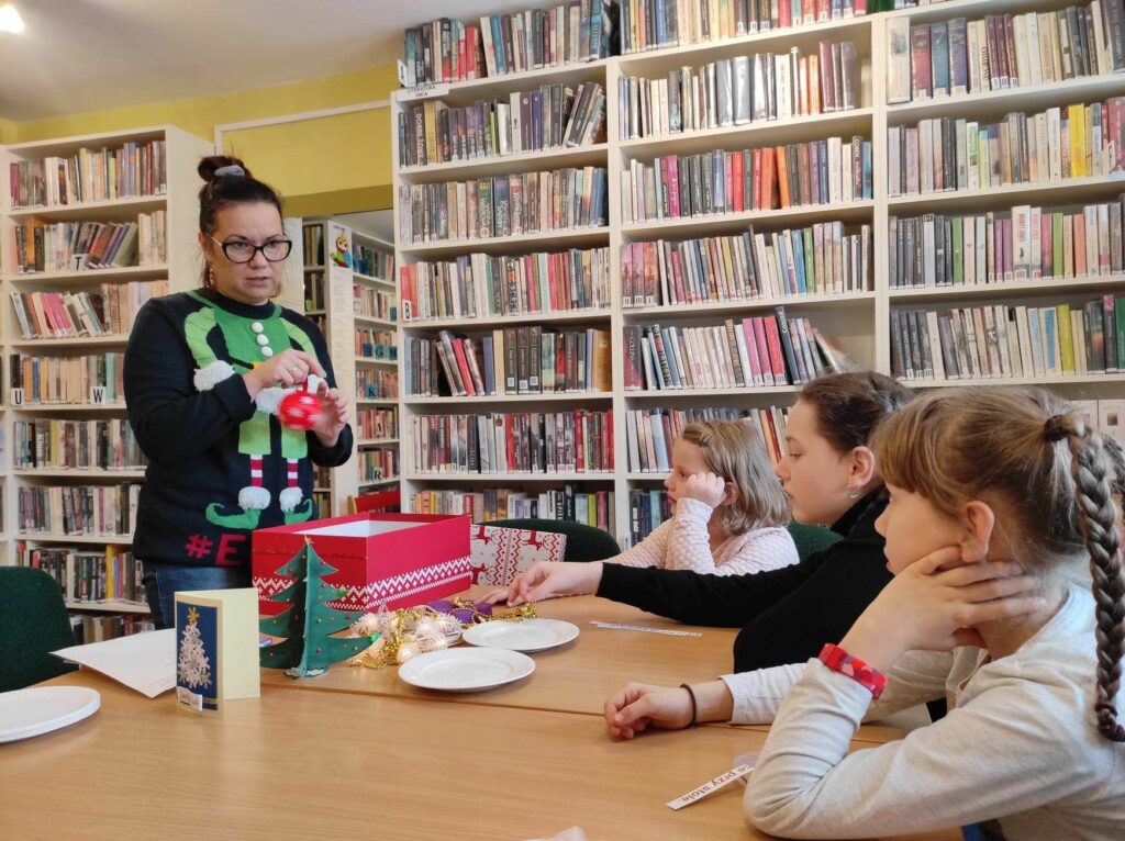 na zdjęciu widać bibliotekarkę w świątecznym swetrze oraz słuchające ją dzieci