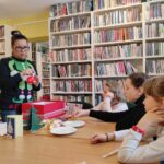 na zdjęciu widać bibliotekarkę w świątecznym swetrze oraz słuchające ją dzieci