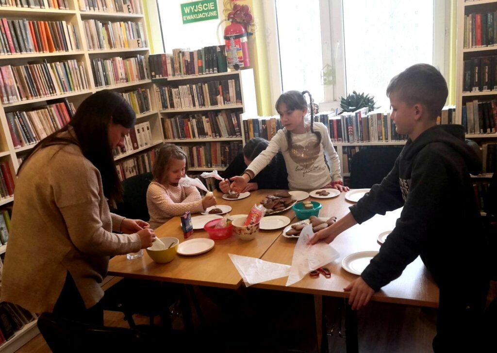 na zdjęciu widać salę biblioteczną oraz stół wokół którego siedzą dzieci i bibliotekarka