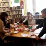 na zdjęciu widać salę biblioteczną oraz stół wokół którego siedzą dzieci i bibliotekarka
