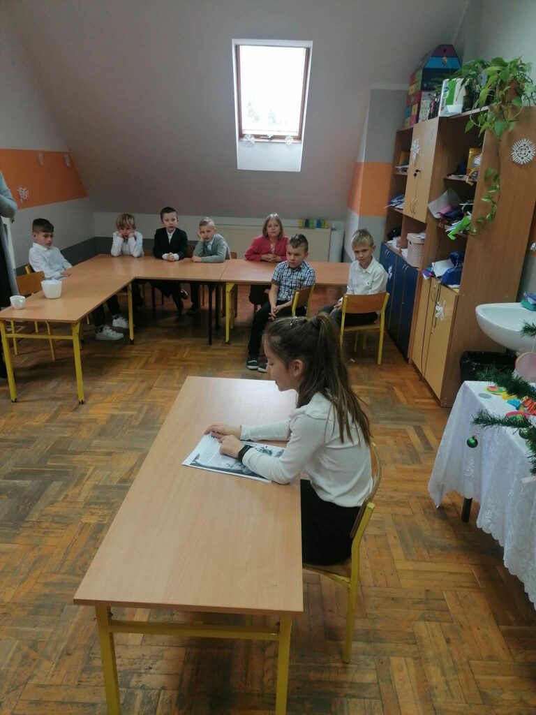 Uczniowie jednej z klas siedzą w ławkach pod oknami, natomiast na środku ich koleżanka czyta w skupieniu wylosowany tekst.