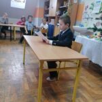 Na środku klasy przy stoliku siedzi ubrany w elegancki garnitur chłopczyk i czyta, starając się robić to jak najładniej, wylosowaną przez siebie historyjkę.