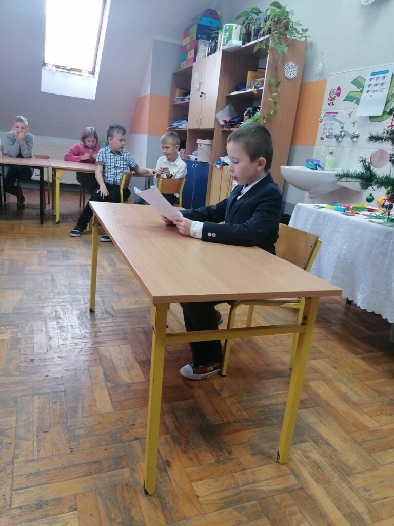 Na środku klasy przy stoliku siedzi ubrany w elegancki garnitur chłopczyk i czyta, starając się robić to jak najładniej, wylosowaną przez siebie historyjkę.