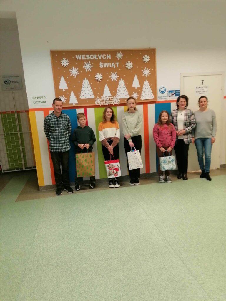 Na środku holu szkolnego mając za plecami gazetkę z zimowym napisem „Wesołych Świąt” stoją uśmiechnięci zwycięzcy konkursów, a w rękach trzymają swoje torby z nagrodami. Obok nich stoi Pani Dyrektor, Pani Bibliotekarka Szkolna oraz Pan Bibliotekarz z Filii w Bereście