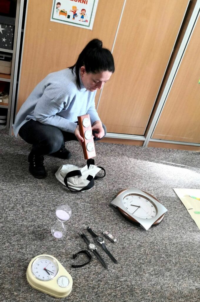 na zdjęciu widać kobietę kucającą na popielatej wykładzinie pokazującą różnego rodzaju zegary.