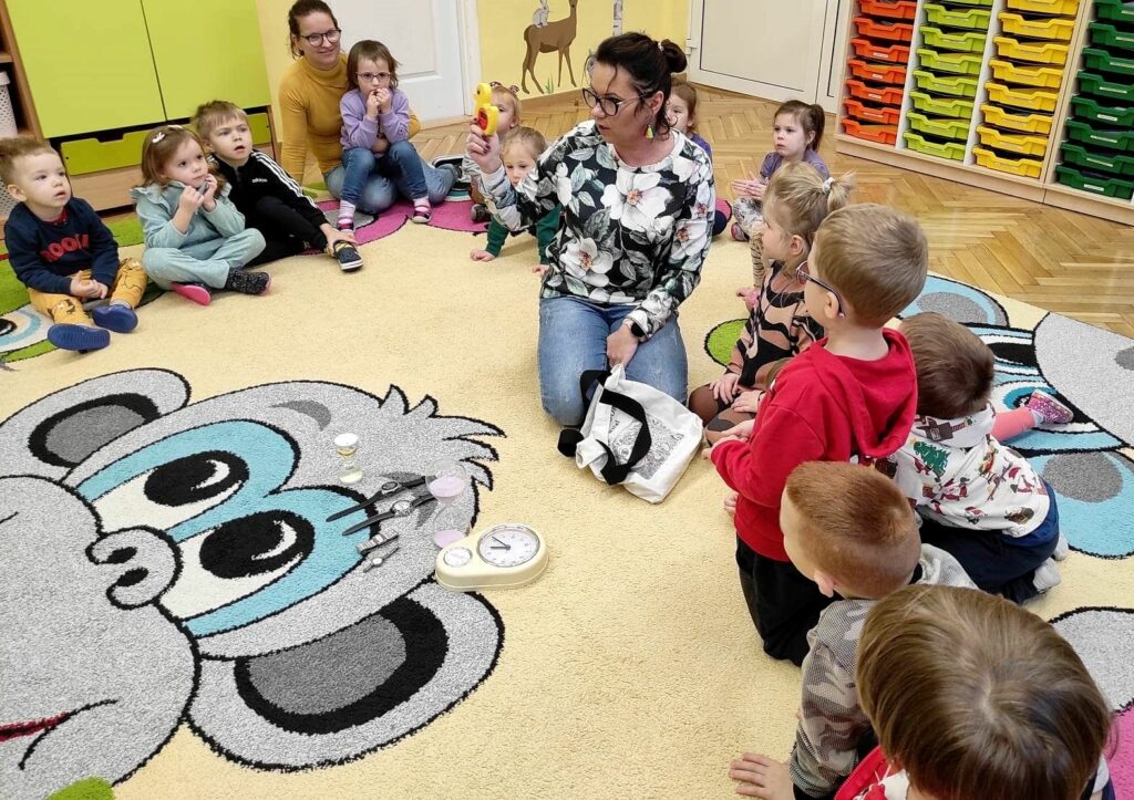 Na zdjęciu widać salę przedszkolna i dzieci na kolorowym dywanie, na którym rozłożono rożnego rodzaju zegary.
