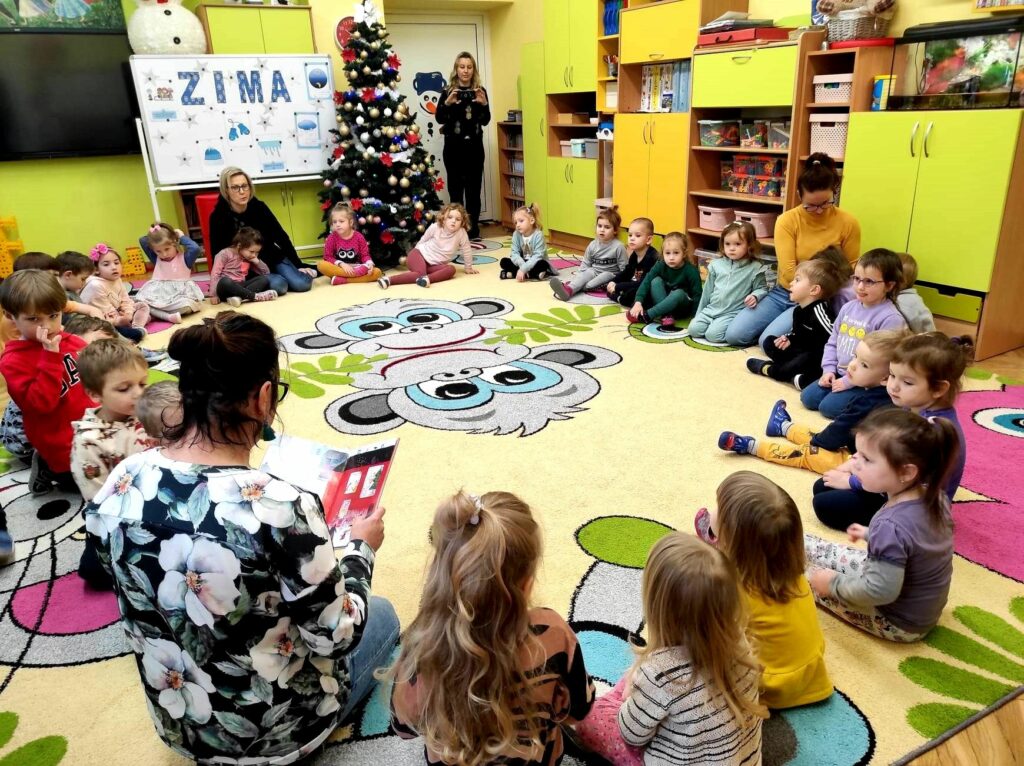 Na zdjęciu widać salę przedszkolną oraz dzieci siedzące na dywanie słuchające czytającej im bibliotekarki